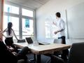 Das Bild zeigt eine Meeting-Situation. Zwei Menschen sitzen am Tisch, eine Person steht daneben und eine weitere stehende Person zeigt etwas an einem Whiteboard.