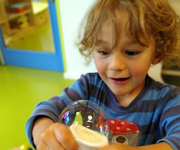 Das Foto zeigt ein etwa 5 Jahre altes Kind, das mit einer selbstgemachten Seifenblasenmaschine experimentiert. Es sieht sehr fasziniert aus.