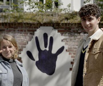 Auf dem Bild sind die zwei Hauptdarsteller:innen des Handabdruck-Films zu sehen, wie sie ihre Hände in die Kamera halten. Hinter ihnen ist ein Handabdruck auf der Mauer zu sehen.