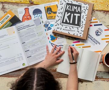 eine Person sitzt an einem Tisch und schreibt etwas in ein Buch, vor ihr viele Materialien aus dem Klima-Kit von Greenpeace