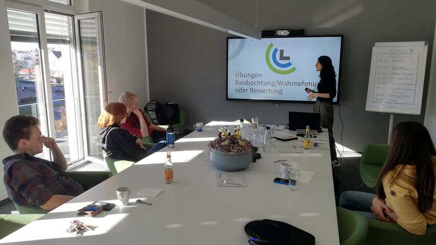 Mehrere Menschen sitzen um einen Tisch. Am Ende des Tischs steht eine Person, die einen Vortrag hält.