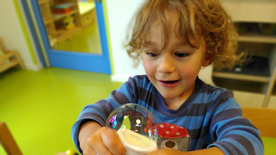 Das Foto zeigt ein etwa 5 Jahre altes Kind, das mit einer selbstgemachten Seifenblasenmaschine experimentiert. Es sieht sehr fasziniert aus.