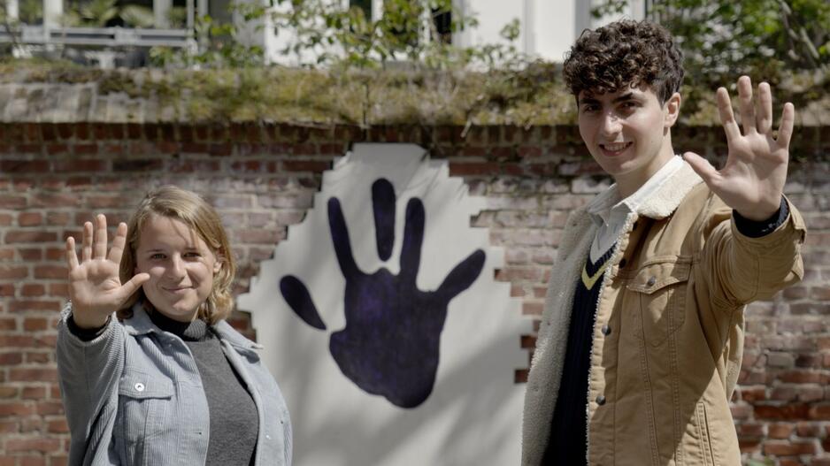 Auf dem Bild sind die zwei Hauptdarsteller:innen des Handabdruck-Films zu sehen, wie sie ihre Hände in die Kamera halten. Hinter ihnen ist ein Handabdruck auf der Mauer zu sehen.