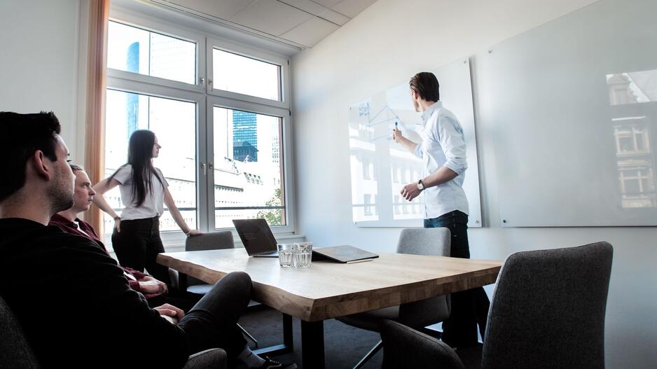 Das Bild zeigt eine Meeting-Situation. Zwei Menschen sitzen am Tisch, eine Person steht daneben und eine weitere stehende Person zeigt etwas an einem Whiteboard.