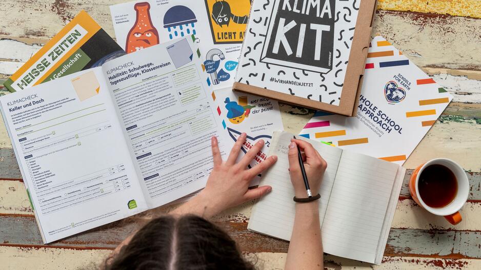 eine Person sitzt an einem Tisch und schreibt etwas in ein Buch, vor ihr viele Materialien aus dem Klima-Kit von Greenpeace