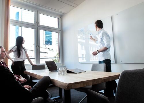 Das Bild zeigt eine Meeting-Situation. Zwei Menschen sitzen am Tisch, eine Person steht daneben und eine weitere stehende Person zeigt etwas an einem Whiteboard.