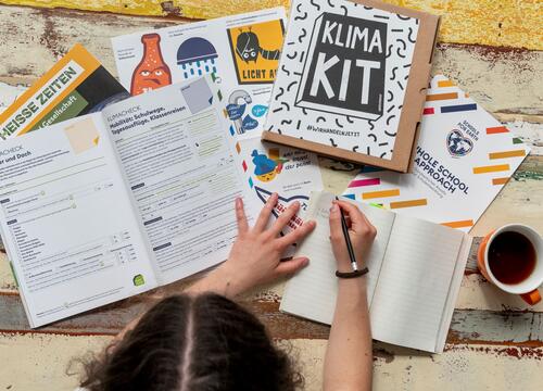 eine Person sitzt an einem Tisch und schreibt etwas in ein Buch, vor ihr viele Materialien aus dem Klima-Kit von Greenpeace