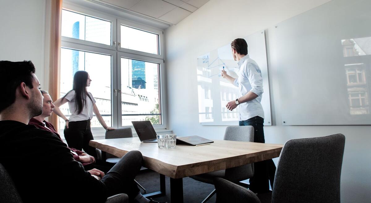Das Bild zeigt eine Meeting-Situation. Zwei Menschen sitzen am Tisch, eine Person steht daneben und eine weitere stehende Person zeigt etwas an einem Whiteboard.